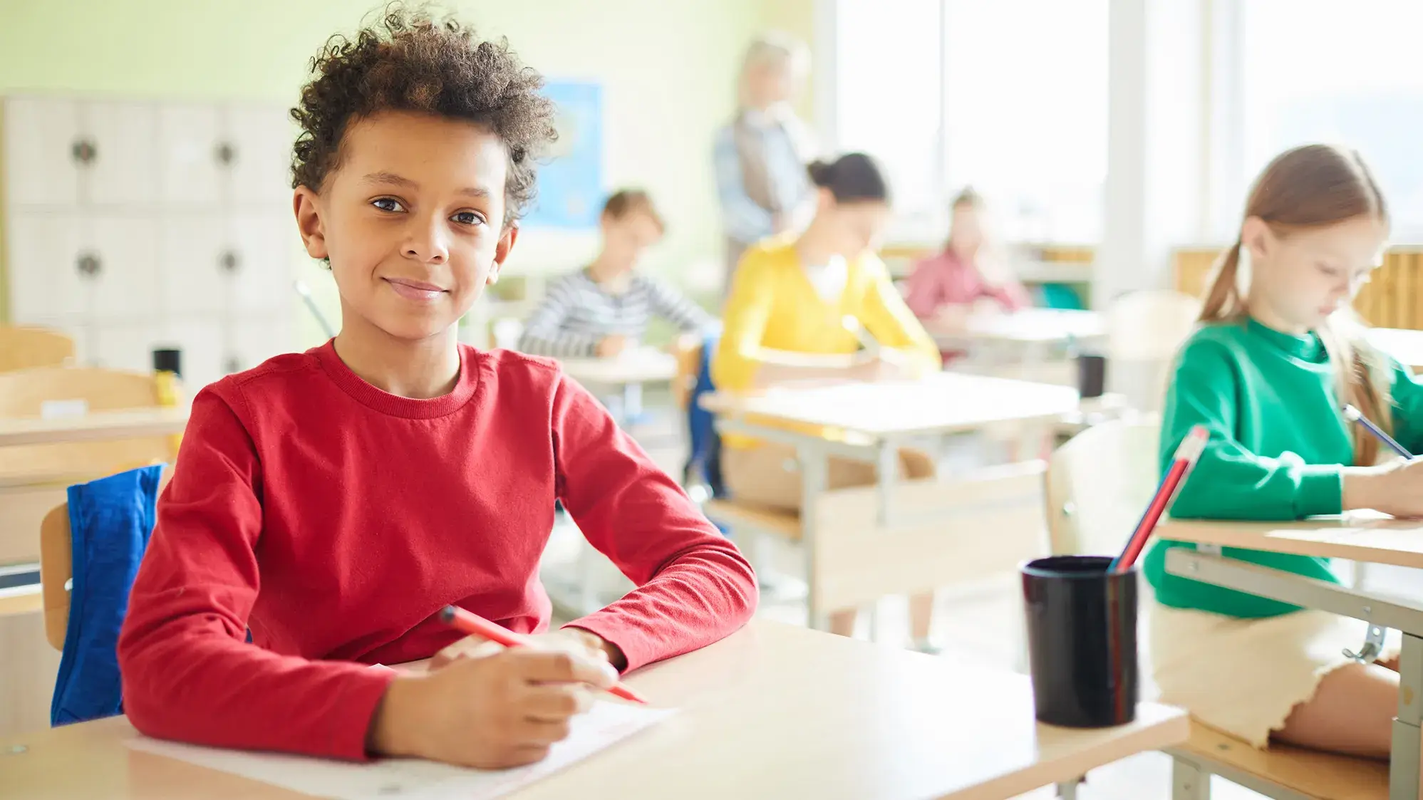 young student smiles as he sits at his desk in a classroom setting